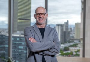 Retrato corporativo en plano medio de Gilberto Garza, Vicepresidente Regional de Ventas de Zendesk Latam, posando sonriente con los brazos cruzados. Viste un saco formal gris claro sobre una playera negra y usa lentes de armazón carey. Se ubica en una oficina moderna frente a un gran ventanal de piso a techo que muestra una vista panorámica difuminada del horizonte urbano de una ciudad con rascacielos bajo un cielo nublado.