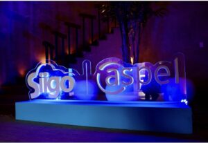 Fotografía en plano general con iluminación de acento en tonalidades azules y violetas de un tótem publicitario volumétrico de acrílico transparente colocado en el suelo. El letrero luminoso muestra los logotipos calados en color blanco de "Siigo | aspel" montados sobre una base rectangular blanca sólida. Al fondo, en la penumbra, se aprecia una planta decorativa de hojas largas y una estructura de escaleras de madera con barandales negros contra un muro rústico.