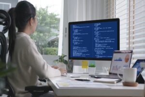 Una mujer de espaldas con anteojos y el cabello recogido en una coleta trabaja frente a su escritorio en una oficina luminosa. Utiliza un monitor de computadora principal que despliega líneas de código de programación sobre un fondo azul, mientras que a la derecha se observa una computadora portátil que muestra gráficos financieros de barras y pasteles junto a una taza de café.