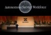 Zendesk redefine IA enterprise con agentes autónomos Fotografía de plano general que captura una conferencia en un gran escenario teatral con piso negro brillante. En el centro, un conferencista varón con vestimenta oscura habla gesticulando con las manos abiertas, parándose frente a una monumental mampara de madera clara con el logotipo y nombre de "Zendesk" en color negro, flanqueada por jardineras con plantas verdes. En la pantalla gigante superior de fondo negro resalta el título luminoso "Autonomous Service Workforce" debajo de un gran gráfico circular formado por miles de partículas blancas brillantes.