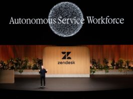Zendesk redefine IA enterprise con agentes autónomos Fotografía de plano general que captura una conferencia en un gran escenario teatral con piso negro brillante. En el centro, un conferencista varón con vestimenta oscura habla gesticulando con las manos abiertas, parándose frente a una monumental mampara de madera clara con el logotipo y nombre de "Zendesk" en color negro, flanqueada por jardineras con plantas verdes. En la pantalla gigante superior de fondo negro resalta el título luminoso "Autonomous Service Workforce" debajo de un gran gráfico circular formado por miles de partículas blancas brillantes.