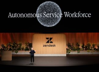 Zendesk redefine IA enterprise con agentes autónomos Fotografía de plano general que captura una conferencia en un gran escenario teatral con piso negro brillante. En el centro, un conferencista varón con vestimenta oscura habla gesticulando con las manos abiertas, parándose frente a una monumental mampara de madera clara con el logotipo y nombre de "Zendesk" en color negro, flanqueada por jardineras con plantas verdes. En la pantalla gigante superior de fondo negro resalta el título luminoso "Autonomous Service Workforce" debajo de un gran gráfico circular formado por miles de partículas blancas brillantes.