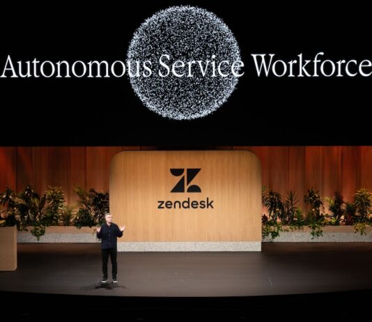 Zendesk redefine IA enterprise con agentes autónomos Fotografía de plano general que captura una conferencia en un gran escenario teatral con piso negro brillante. En el centro, un conferencista varón con vestimenta oscura habla gesticulando con las manos abiertas, parándose frente a una monumental mampara de madera clara con el logotipo y nombre de "Zendesk" en color negro, flanqueada por jardineras con plantas verdes. En la pantalla gigante superior de fondo negro resalta el título luminoso "Autonomous Service Workforce" debajo de un gran gráfico circular formado por miles de partículas blancas brillantes.