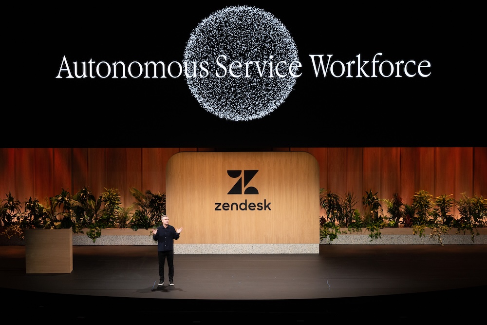 Zendesk presenta Autonomous Service Workforce para redefinir la automatización corporativa Fotografía de plano general que captura una conferencia en un gran escenario teatral con piso negro brillante. En el centro, un conferencista varón con vestimenta oscura habla gesticulando con las manos abiertas, parándose frente a una monumental mampara de madera clara con el logotipo y nombre de