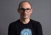 Dynatrace visualiza una nueva etapa para las operaciones autónomas impulsadas por IA Retrato de estudio en plano medio y de frente de Bernd Greifeneder, fundador y CTO de Dynatrace. Viste una playera negra que tiene estampado en el pecho un gráfico circular abstracto de color azul brillante con un cubo en su interior. Luce el cabello muy corto, usa lentes de armazón delgado de color oscuro y sonríe ligeramente con la mirada directa a la cámara. Se encuentra posando frente a un fondo gris neutro, liso y con iluminación tenue.