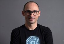 Dynatrace visualiza una nueva etapa para las operaciones autónomas impulsadas por IA Retrato de estudio en plano medio y de frente de Bernd Greifeneder, fundador y CTO de Dynatrace. Viste una playera negra que tiene estampado en el pecho un gráfico circular abstracto de color azul brillante con un cubo en su interior. Luce el cabello muy corto, usa lentes de armazón delgado de color oscuro y sonríe ligeramente con la mirada directa a la cámara. Se encuentra posando frente a un fondo gris neutro, liso y con iluminación tenue.