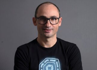 Dynatrace visualiza una nueva etapa para las operaciones autónomas impulsadas por IA Retrato de estudio en plano medio y de frente de Bernd Greifeneder, fundador y CTO de Dynatrace. Viste una playera negra que tiene estampado en el pecho un gráfico circular abstracto de color azul brillante con un cubo en su interior. Luce el cabello muy corto, usa lentes de armazón delgado de color oscuro y sonríe ligeramente con la mirada directa a la cámara. Se encuentra posando frente a un fondo gris neutro, liso y con iluminación tenue.
