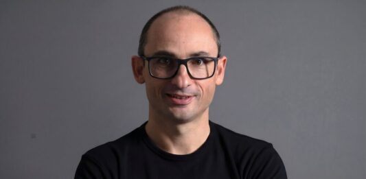 Dynatrace visualiza una nueva etapa para las operaciones autónomas impulsadas por IA Retrato de estudio en plano medio y de frente de Bernd Greifeneder, fundador y CTO de Dynatrace. Viste una playera negra que tiene estampado en el pecho un gráfico circular abstracto de color azul brillante con un cubo en su interior. Luce el cabello muy corto, usa lentes de armazón delgado de color oscuro y sonríe ligeramente con la mirada directa a la cámara. Se encuentra posando frente a un fondo gris neutro, liso y con iluminación tenue.