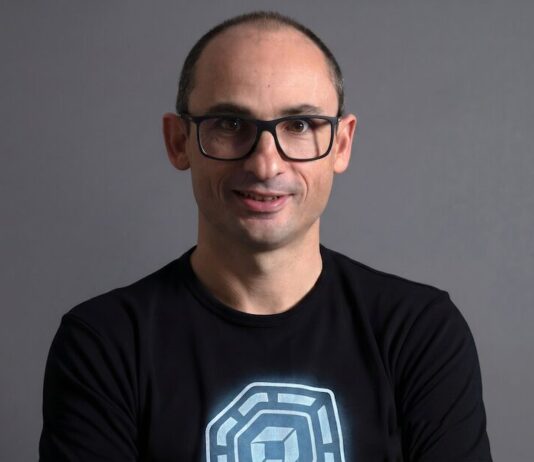 Dynatrace visualiza una nueva etapa para las operaciones autónomas impulsadas por IA Retrato de estudio en plano medio y de frente de Bernd Greifeneder, fundador y CTO de Dynatrace. Viste una playera negra que tiene estampado en el pecho un gráfico circular abstracto de color azul brillante con un cubo en su interior. Luce el cabello muy corto, usa lentes de armazón delgado de color oscuro y sonríe ligeramente con la mirada directa a la cámara. Se encuentra posando frente a un fondo gris neutro, liso y con iluminación tenue.