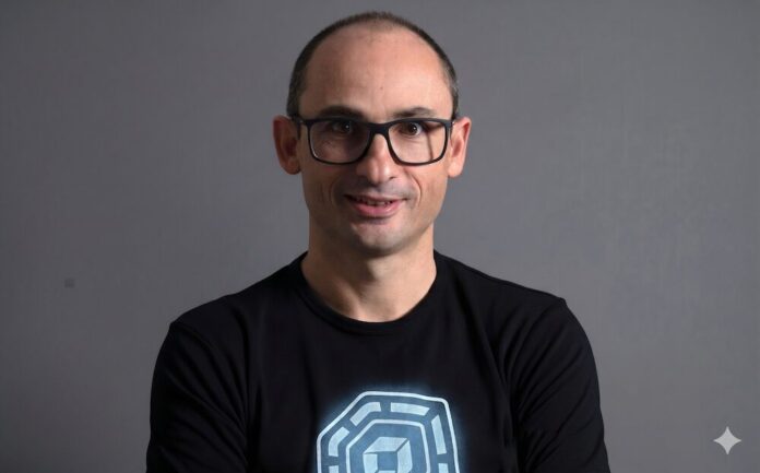 Retrato de estudio en plano medio y de frente de Bernd Greifeneder, fundador y CTO de Dynatrace. Viste una playera negra que tiene estampado en el pecho un gráfico circular abstracto de color azul brillante con un cubo en su interior. Luce el cabello muy corto, usa lentes de armazón delgado de color oscuro y sonríe ligeramente con la mirada directa a la cámara. Se encuentra posando frente a un fondo gris neutro, liso y con iluminación tenue.