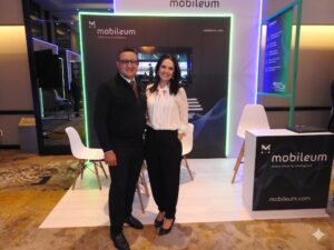 Fotografía en plano general medio de una pareja de colaboradores de Mobileum posando sonrientes de frente en el stand corporativo de la empresa. El hombre, ubicado a la izquierda, tiene tez clara, cabello corto oscuro y anteojos de armazón negro, y viste un chaleco ejecutivo negro sobre camisa blanca con corbata roja y pantalón oscuro. La mujer, a la derecha, de tez clara y cabello largo oscuro, viste una blusa blanca elegante de cuello de lazo con mangas largas y un pantalón de vestir negro de talle alto. Al fondo, la mampara principal del stand muestra un panel negro con iluminación de luces LED de color verde fosforescente y el logotipo blanco de "Mobileum - Action driven by intelligence".