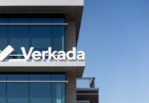 Verkada coloca a México al frente de su estrategia de crecimiento en América Latina Fotografía en ángulo contrapicado del exterior de un edificio corporativo moderno con fachadas de cristal azul reflectante bajo un cielo despejado. En la cornisa de la terraza superior destaca el letrero comercial blanco en relieve con la tipografía oficial de Verkada junto a su logotipo icónico en forma de marca de verificación o doble chevron estilizado.