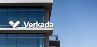 Verkada coloca a México al frente de su estrategia de crecimiento en América Latina Fotografía en ángulo contrapicado del exterior de un edificio corporativo moderno con fachadas de cristal azul reflectante bajo un cielo despejado. En la cornisa de la terraza superior destaca el letrero comercial blanco en relieve con la tipografía oficial de Verkada junto a su logotipo icónico en forma de marca de verificación o doble chevron estilizado.