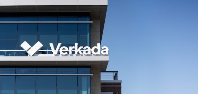 Fotografía en ángulo contrapicado del exterior de un edificio corporativo moderno con fachadas de cristal azul reflectante bajo un cielo despejado. En la cornisa de la terraza superior destaca el letrero comercial blanco en relieve con la tipografía oficial de Verkada junto a su logotipo icónico en forma de marca de verificación o doble chevron estilizado.