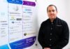 Intercompras: El eCommerce empresarial redefine la forma en que las organizaciones compran tecnología Retrato corporativo de plano medio que muestra a Eduardo Noriega, CEO de Intercompras, vistiendo una camisa negra formal con un sutil logotipo bordado en el pecho. Posa de tres cuartos hacia la izquierda, sonriendo levemente frente a una pared blanca donde cuelga un gran panel acrílico transparente. El panel despliega una línea de tiempo corporativa dividida en secciones cronológicas con logotipos de marcas como Cisco, Syscom, Unicom y Grupo Decme, detallando hitos de infraestructura, volumen de productos, cantidad de clientes y una sección inferior titulada "Rumbo al 2030: El futuro que estamos construyendo".
