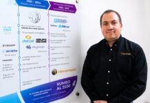 Intercompras: El eCommerce empresarial redefine la forma en que las organizaciones compran tecnología Retrato corporativo de plano medio que muestra a Eduardo Noriega, CEO de Intercompras, vistiendo una camisa negra formal con un sutil logotipo bordado en el pecho. Posa de tres cuartos hacia la izquierda, sonriendo levemente frente a una pared blanca donde cuelga un gran panel acrílico transparente. El panel despliega una línea de tiempo corporativa dividida en secciones cronológicas con logotipos de marcas como Cisco, Syscom, Unicom y Grupo Decme, detallando hitos de infraestructura, volumen de productos, cantidad de clientes y una sección inferior titulada "Rumbo al 2030: El futuro que estamos construyendo".