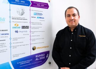 Intercompras: El eCommerce empresarial redefine la forma en que las organizaciones compran tecnología Retrato corporativo de plano medio que muestra a Eduardo Noriega, CEO de Intercompras, vistiendo una camisa negra formal con un sutil logotipo bordado en el pecho. Posa de tres cuartos hacia la izquierda, sonriendo levemente frente a una pared blanca donde cuelga un gran panel acrílico transparente. El panel despliega una línea de tiempo corporativa dividida en secciones cronológicas con logotipos de marcas como Cisco, Syscom, Unicom y Grupo Decme, detallando hitos de infraestructura, volumen de productos, cantidad de clientes y una sección inferior titulada "Rumbo al 2030: El futuro que estamos construyendo".