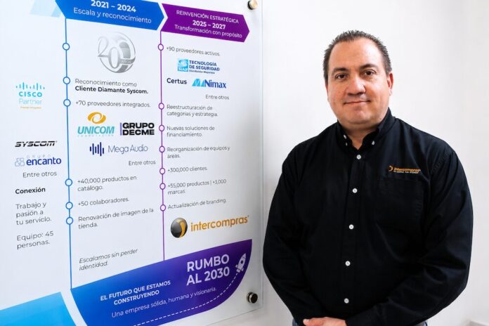 Retrato corporativo de plano medio que muestra a Eduardo Noriega, CEO de Intercompras, vistiendo una camisa negra formal con un sutil logotipo bordado en el pecho. Posa de tres cuartos hacia la izquierda, sonriendo levemente frente a una pared blanca donde cuelga un gran panel acrílico transparente. El panel despliega una línea de tiempo corporativa dividida en secciones cronológicas con logotipos de marcas como Cisco, Syscom, Unicom y Grupo Decme, detallando hitos de infraestructura, volumen de productos, cantidad de clientes y una sección inferior titulada 