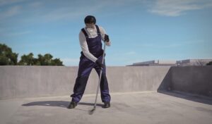 Fotografía en plano medio largo que muestra a un aplicador de espaldas inclinadas sobre la superficie plana de una azotea de concreto en un día despejado con cielo azul. El trabajador utiliza un overol de protección azul marino sobre una playera blanca de manga larga, guantes negros, lentes de seguridad y una mascarilla desechable contra polvo. Con ambas manos sostiene firmemente el mango largo de un rodillo o jalador de felpa con el que extiende una capa uniforme de recubrimiento líquido sobre el piso gris. Al fondo se distinguen el borde perimetral de la azotea, algunos árboles verdes dispersos a la izquierda y el contorno de un edificio industrial a la derecha.