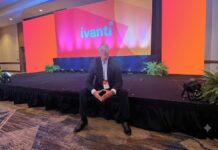 Ivanti impulsa operaciones autónomas de TI y ciberseguridad con IA en América Latina Fotografía en plano general medio que muestra a Dennis Kozak sentado al borde de un escenario alfombrado en un salón de conferencias durante un evento corporativo. Detrás de él se alza una gran pantalla gigante iluminada en tonalidades brillantes de naranja, rosa y morado que despliega al centro el logotipo oficial de "ivanti" en letras turquesas. El directivo viste un traje oscuro con camisa clara y un gafete oficial colgado del cuello, manteniendo los dedos de ambas manos entrelazados al frente mientras posa sonriente para la cámara.