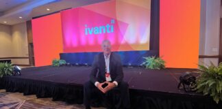 Ivanti impulsa operaciones autónomas de TI y ciberseguridad con IA en América Latina Fotografía en plano general medio que muestra a Dennis Kozak sentado al borde de un escenario alfombrado en un salón de conferencias durante un evento corporativo. Detrás de él se alza una gran pantalla gigante iluminada en tonalidades brillantes de naranja, rosa y morado que despliega al centro el logotipo oficial de "ivanti" en letras turquesas. El directivo viste un traje oscuro con camisa clara y un gafete oficial colgado del cuello, manteniendo los dedos de ambas manos entrelazados al frente mientras posa sonriente para la cámara.