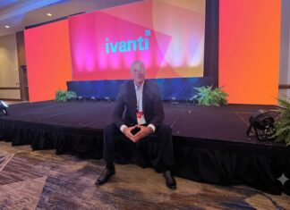 Ivanti impulsa operaciones autónomas de TI y ciberseguridad con IA en América Latina Fotografía en plano general medio que muestra a Dennis Kozak sentado al borde de un escenario alfombrado en un salón de conferencias durante un evento corporativo. Detrás de él se alza una gran pantalla gigante iluminada en tonalidades brillantes de naranja, rosa y morado que despliega al centro el logotipo oficial de "ivanti" en letras turquesas. El directivo viste un traje oscuro con camisa clara y un gafete oficial colgado del cuello, manteniendo los dedos de ambas manos entrelazados al frente mientras posa sonriente para la cámara.