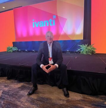 Ivanti impulsa operaciones autónomas de TI y ciberseguridad con IA en América Latina Fotografía en plano general medio que muestra a Dennis Kozak sentado al borde de un escenario alfombrado en un salón de conferencias durante un evento corporativo. Detrás de él se alza una gran pantalla gigante iluminada en tonalidades brillantes de naranja, rosa y morado que despliega al centro el logotipo oficial de "ivanti" en letras turquesas. El directivo viste un traje oscuro con camisa clara y un gafete oficial colgado del cuello, manteniendo los dedos de ambas manos entrelazados al frente mientras posa sonriente para la cámara.
