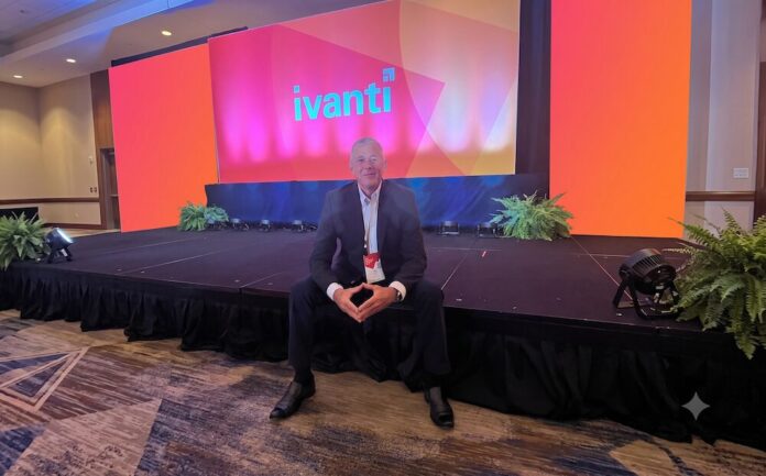Fotografía en plano general medio que muestra a Dennis Kozak sentado al borde de un escenario alfombrado en un salón de conferencias durante un evento corporativo. Detrás de él se alza una gran pantalla gigante iluminada en tonalidades brillantes de naranja, rosa y morado que despliega al centro el logotipo oficial de 