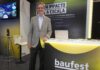 Inteligencia artificial empresarial: Baufest apuesta por valor Fotografía en plano medio que muestra a Luis Battilana, Country Manager de Baufest, posando de frente con una sonrisa amable en un espacio de exposición. El ejecutivo luce un saco sport gris claro sobre una camisa azul claro y pantalón de vestir oscuro, manteniendo su mano izquierda apoyada sobre un mostrador publicitario negro con bordes iluminados en amarillo cian que exhibe el logotipo de baufest evolving business together. Detrás de él resalta una mampara oscura con la inscripción en letras blancas gigantes IA CON IMPACTO REAL A ESCALA, mientras que a la izquierda se observa una pantalla de televisión encendida con imágenes de conferencistas.