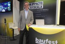 Inteligencia artificial empresarial: Baufest apuesta por valor Fotografía en plano medio que muestra a Luis Battilana, Country Manager de Baufest, posando de frente con una sonrisa amable en un espacio de exposición. El ejecutivo luce un saco sport gris claro sobre una camisa azul claro y pantalón de vestir oscuro, manteniendo su mano izquierda apoyada sobre un mostrador publicitario negro con bordes iluminados en amarillo cian que exhibe el logotipo de baufest evolving business together. Detrás de él resalta una mampara oscura con la inscripción en letras blancas gigantes IA CON IMPACTO REAL A ESCALA, mientras que a la izquierda se observa una pantalla de televisión encendida con imágenes de conferencistas.