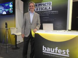 Inteligencia artificial empresarial: Baufest apuesta por valor Fotografía en plano medio que muestra a Luis Battilana, Country Manager de Baufest, posando de frente con una sonrisa amable en un espacio de exposición. El ejecutivo luce un saco sport gris claro sobre una camisa azul claro y pantalón de vestir oscuro, manteniendo su mano izquierda apoyada sobre un mostrador publicitario negro con bordes iluminados en amarillo cian que exhibe el logotipo de baufest evolving business together. Detrás de él resalta una mampara oscura con la inscripción en letras blancas gigantes IA CON IMPACTO REAL A ESCALA, mientras que a la izquierda se observa una pantalla de televisión encendida con imágenes de conferencistas.