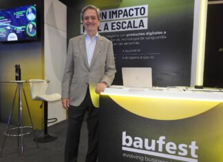 Inteligencia artificial empresarial: Baufest apuesta por valor Fotografía en plano medio que muestra a Luis Battilana, Country Manager de Baufest, posando de frente con una sonrisa amable en un espacio de exposición. El ejecutivo luce un saco sport gris claro sobre una camisa azul claro y pantalón de vestir oscuro, manteniendo su mano izquierda apoyada sobre un mostrador publicitario negro con bordes iluminados en amarillo cian que exhibe el logotipo de baufest evolving business together. Detrás de él resalta una mampara oscura con la inscripción en letras blancas gigantes IA CON IMPACTO REAL A ESCALA, mientras que a la izquierda se observa una pantalla de televisión encendida con imágenes de conferencistas.