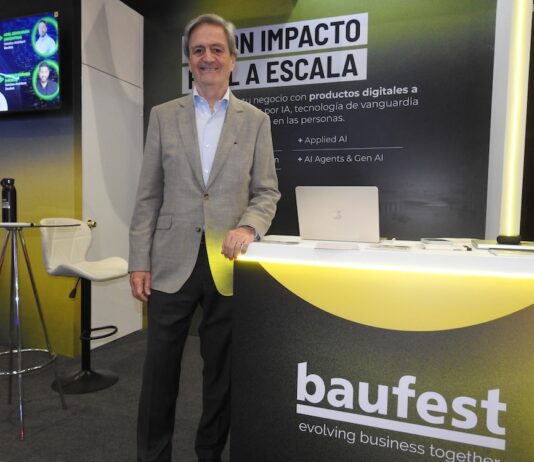 Inteligencia artificial empresarial: Baufest apuesta por valor Fotografía en plano medio que muestra a Luis Battilana, Country Manager de Baufest, posando de frente con una sonrisa amable en un espacio de exposición. El ejecutivo luce un saco sport gris claro sobre una camisa azul claro y pantalón de vestir oscuro, manteniendo su mano izquierda apoyada sobre un mostrador publicitario negro con bordes iluminados en amarillo cian que exhibe el logotipo de baufest evolving business together. Detrás de él resalta una mampara oscura con la inscripción en letras blancas gigantes IA CON IMPACTO REAL A ESCALA, mientras que a la izquierda se observa una pantalla de televisión encendida con imágenes de conferencistas.