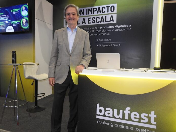 Fotografía en plano medio que muestra a Luis Battilana, Country Manager de Baufest, posando de frente con una sonrisa amable en un espacio de exposición. El ejecutivo luce un saco sport gris claro sobre una camisa azul claro y pantalón de vestir oscuro, manteniendo su mano izquierda apoyada sobre un mostrador publicitario negro con bordes iluminados en amarillo cian que exhibe el logotipo de baufest evolving business together. Detrás de él resalta una mampara oscura con la inscripción en letras blancas gigantes IA CON IMPACTO REAL A ESCALA, mientras que a la izquierda se observa una pantalla de televisión encendida con imágenes de conferencistas.