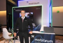 Mobileum y la evolución del ecosistema telecom en Latinoamérica Fotografía de frente en plano medio largo que muestra a Omar de Jesús Díaz, director comercial para el norte de Latinoamérica de Mobileum, de pie y apoyando su mano izquierda sobre un mostrador publicitario negro con la leyenda "Mobileum - Action driven by intelligence". El directivo, de complexión media, tez clara y anteojos de armazón transparente, sonríe formalmente vistiendo una chamarra ejecutiva negra con cierre sobre una camisa blanca, corbata roja texturizada y pantalón de vestir oscuro. Detrás de él, el stand cuenta con iluminación decorativa LED azul y verde alrededor de una pantalla de televisión empotrada.