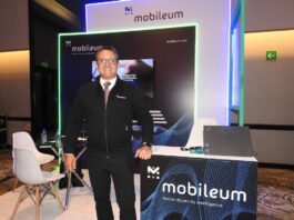 Mobileum y la evolución del ecosistema telecom en Latinoamérica Fotografía de frente en plano medio largo que muestra a Omar de Jesús Díaz, director comercial para el norte de Latinoamérica de Mobileum, de pie y apoyando su mano izquierda sobre un mostrador publicitario negro con la leyenda "Mobileum - Action driven by intelligence". El directivo, de complexión media, tez clara y anteojos de armazón transparente, sonríe formalmente vistiendo una chamarra ejecutiva negra con cierre sobre una camisa blanca, corbata roja texturizada y pantalón de vestir oscuro. Detrás de él, el stand cuenta con iluminación decorativa LED azul y verde alrededor de una pantalla de televisión empotrada.