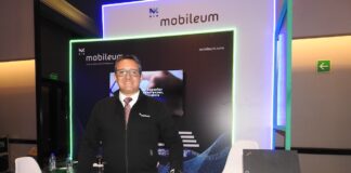 Mobileum y la evolución del ecosistema telecom en Latinoamérica Fotografía de frente en plano medio largo que muestra a Omar de Jesús Díaz, director comercial para el norte de Latinoamérica de Mobileum, de pie y apoyando su mano izquierda sobre un mostrador publicitario negro con la leyenda "Mobileum - Action driven by intelligence". El directivo, de complexión media, tez clara y anteojos de armazón transparente, sonríe formalmente vistiendo una chamarra ejecutiva negra con cierre sobre una camisa blanca, corbata roja texturizada y pantalón de vestir oscuro. Detrás de él, el stand cuenta con iluminación decorativa LED azul y verde alrededor de una pantalla de televisión empotrada.