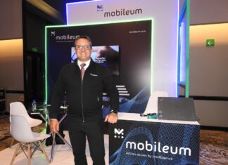 Mobileum y la evolución del ecosistema telecom en Latinoamérica Fotografía de frente en plano medio largo que muestra a Omar de Jesús Díaz, director comercial para el norte de Latinoamérica de Mobileum, de pie y apoyando su mano izquierda sobre un mostrador publicitario negro con la leyenda "Mobileum - Action driven by intelligence". El directivo, de complexión media, tez clara y anteojos de armazón transparente, sonríe formalmente vistiendo una chamarra ejecutiva negra con cierre sobre una camisa blanca, corbata roja texturizada y pantalón de vestir oscuro. Detrás de él, el stand cuenta con iluminación decorativa LED azul y verde alrededor de una pantalla de televisión empotrada.