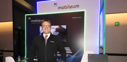 Mobileum y la evolución del ecosistema telecom en Latinoamérica Fotografía de frente en plano medio largo que muestra a Omar de Jesús Díaz, director comercial para el norte de Latinoamérica de Mobileum, de pie y apoyando su mano izquierda sobre un mostrador publicitario negro con la leyenda "Mobileum - Action driven by intelligence". El directivo, de complexión media, tez clara y anteojos de armazón transparente, sonríe formalmente vistiendo una chamarra ejecutiva negra con cierre sobre una camisa blanca, corbata roja texturizada y pantalón de vestir oscuro. Detrás de él, el stand cuenta con iluminación decorativa LED azul y verde alrededor de una pantalla de televisión empotrada.