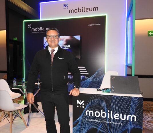 Mobileum y la evolución del ecosistema telecom en Latinoamérica Fotografía de frente en plano medio largo que muestra a Omar de Jesús Díaz, director comercial para el norte de Latinoamérica de Mobileum, de pie y apoyando su mano izquierda sobre un mostrador publicitario negro con la leyenda "Mobileum - Action driven by intelligence". El directivo, de complexión media, tez clara y anteojos de armazón transparente, sonríe formalmente vistiendo una chamarra ejecutiva negra con cierre sobre una camisa blanca, corbata roja texturizada y pantalón de vestir oscuro. Detrás de él, el stand cuenta con iluminación decorativa LED azul y verde alrededor de una pantalla de televisión empotrada.