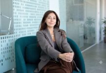 Mambu acelera la banca con IA para reducir costos e impulsar la innovación financiera Fotografía en plano medio de Paula Neira, directora de ventas de Mambu para América Latina, sentada cómodamente en un moderno sillón ejecutivo de color verde tejavana o turquesa oscuro dentro de un entorno de oficina iluminado. La directiva, de cabello largo oscuro y tez clara, viste un blazer casual a cuadros finos estilo pata de gallo en tonos marrón y gris, combinado con una blusa interior oscura, pantalón café y un collar de cadena delgada con un dije circular plateado. Mantiene una sonrisa sutil frente a la cámara con las manos entrelazadas sobre su regazo, mientras al fondo se aprecian mamparas de cristal con patrones esmerilados y una planta decorativa difuminada.