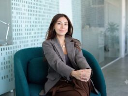 Mambu acelera la banca con IA para reducir costos e impulsar la innovación financiera Fotografía en plano medio de Paula Neira, directora de ventas de Mambu para América Latina, sentada cómodamente en un moderno sillón ejecutivo de color verde tejavana o turquesa oscuro dentro de un entorno de oficina iluminado. La directiva, de cabello largo oscuro y tez clara, viste un blazer casual a cuadros finos estilo pata de gallo en tonos marrón y gris, combinado con una blusa interior oscura, pantalón café y un collar de cadena delgada con un dije circular plateado. Mantiene una sonrisa sutil frente a la cámara con las manos entrelazadas sobre su regazo, mientras al fondo se aprecian mamparas de cristal con patrones esmerilados y una planta decorativa difuminada.