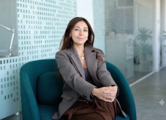 Mambu acelera la banca con IA para reducir costos e impulsar la innovación financiera Fotografía en plano medio de Paula Neira, directora de ventas de Mambu para América Latina, sentada cómodamente en un moderno sillón ejecutivo de color verde tejavana o turquesa oscuro dentro de un entorno de oficina iluminado. La directiva, de cabello largo oscuro y tez clara, viste un blazer casual a cuadros finos estilo pata de gallo en tonos marrón y gris, combinado con una blusa interior oscura, pantalón café y un collar de cadena delgada con un dije circular plateado. Mantiene una sonrisa sutil frente a la cámara con las manos entrelazadas sobre su regazo, mientras al fondo se aprecian mamparas de cristal con patrones esmerilados y una planta decorativa difuminada.