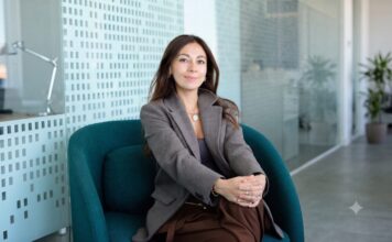 Mambu acelera la banca con IA para reducir costos e impulsar la innovación financiera Fotografía en plano medio de Paula Neira, directora de ventas de Mambu para América Latina, sentada cómodamente en un moderno sillón ejecutivo de color verde tejavana o turquesa oscuro dentro de un entorno de oficina iluminado. La directiva, de cabello largo oscuro y tez clara, viste un blazer casual a cuadros finos estilo pata de gallo en tonos marrón y gris, combinado con una blusa interior oscura, pantalón café y un collar de cadena delgada con un dije circular plateado. Mantiene una sonrisa sutil frente a la cámara con las manos entrelazadas sobre su regazo, mientras al fondo se aprecian mamparas de cristal con patrones esmerilados y una planta decorativa difuminada.