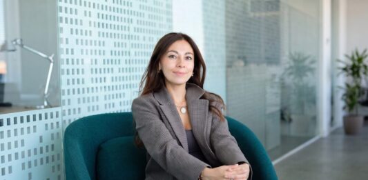 Mambu acelera la banca con IA para reducir costos e impulsar la innovación financiera Fotografía en plano medio de Paula Neira, directora de ventas de Mambu para América Latina, sentada cómodamente en un moderno sillón ejecutivo de color verde tejavana o turquesa oscuro dentro de un entorno de oficina iluminado. La directiva, de cabello largo oscuro y tez clara, viste un blazer casual a cuadros finos estilo pata de gallo en tonos marrón y gris, combinado con una blusa interior oscura, pantalón café y un collar de cadena delgada con un dije circular plateado. Mantiene una sonrisa sutil frente a la cámara con las manos entrelazadas sobre su regazo, mientras al fondo se aprecian mamparas de cristal con patrones esmerilados y una planta decorativa difuminada.