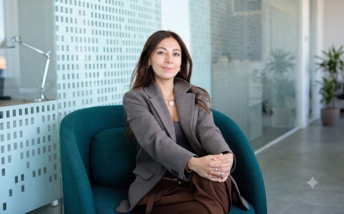Fotografía en plano medio de Paula Neira, directora de ventas de Mambu para América Latina, sentada cómodamente en un moderno sillón ejecutivo de color verde tejavana o turquesa oscuro dentro de un entorno de oficina iluminado. La directiva, de cabello largo oscuro y tez clara, viste un blazer casual a cuadros finos estilo pata de gallo en tonos marrón y gris, combinado con una blusa interior oscura, pantalón café y un collar de cadena delgada con un dije circular plateado. Mantiene una sonrisa sutil frente a la cámara con las manos entrelazadas sobre su regazo, mientras al fondo se aprecian mamparas de cristal con patrones esmerilados y una planta decorativa difuminada.