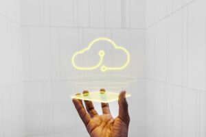 Fotografía en primer plano que muestra una mano sosteniendo una placa de acrílico transparente de la cual emerge un holograma brillante en color amarillo neón con la silueta de una nube interconectada. El fondo blanco minimalista con paneles geométricos resalta la representación conceptual del almacenamiento en la nube y los agentes inteligentes.