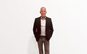 Fotografía de estudio en plano general medio que muestra a Dennis Kozak, CEO de Ivanti, posando de frente sobre un fondo completamente blanco. El ejecutivo, de cabello corto entrecano, viste un saco formal negro sobre una camisa de vestir gris claro de rayas finas y un pantalón casual de tono café claro, manteniendo una postura relajada con ambas manos introducidas de forma parcial en los bolsillos del pantalón.
