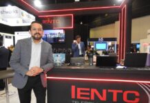 IENTC impulsa la conectividad empresarial para organizaciones que aceleran su transformación digital Fotografía en plano medio largo que captura a Rubén Vargas, manager del segmento corporativo de IENTC, posando de pie junto al módulo de atención de su empresa durante el encuentro de tecnología América Digital 2026. El directivo, con barba corta recortada, viste un saco gris claro con diseño de cuadros finos sobre una camisa formal clara y pantalón oscuro. Se apoya sutilmente en un mostrador negro decorado con tiras de luces LED de neón rojo y el logotipo de "IENTC TELECOMUNICACIONES". Sobre la mesa se aprecian folletos del "11° Congreso IA", computadoras portátiles y termos. Detrás de él, se observa a otro ejecutivo hablando por teléfono en un entorno de exposición dinámico con stands de las marcas freshworks y N5 bajo iluminación de plafón técnica.
