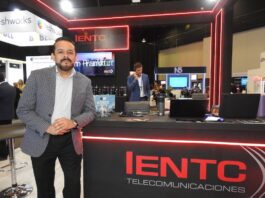 IENTC impulsa la conectividad empresarial para organizaciones que aceleran su transformación digital Fotografía en plano medio largo que captura a Rubén Vargas, manager del segmento corporativo de IENTC, posando de pie junto al módulo de atención de su empresa durante el encuentro de tecnología América Digital 2026. El directivo, con barba corta recortada, viste un saco gris claro con diseño de cuadros finos sobre una camisa formal clara y pantalón oscuro. Se apoya sutilmente en un mostrador negro decorado con tiras de luces LED de neón rojo y el logotipo de "IENTC TELECOMUNICACIONES". Sobre la mesa se aprecian folletos del "11° Congreso IA", computadoras portátiles y termos. Detrás de él, se observa a otro ejecutivo hablando por teléfono en un entorno de exposición dinámico con stands de las marcas freshworks y N5 bajo iluminación de plafón técnica.
